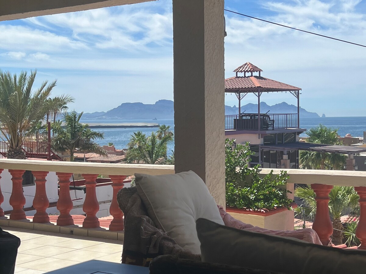 A comfortable outdoor seating area offers a view of the ocean and distant mountains. Palm trees and other greenery surround the space, while a covered structure with a red roof is visible nearby. The scene is framed by a light-colored railing and tiled floor.