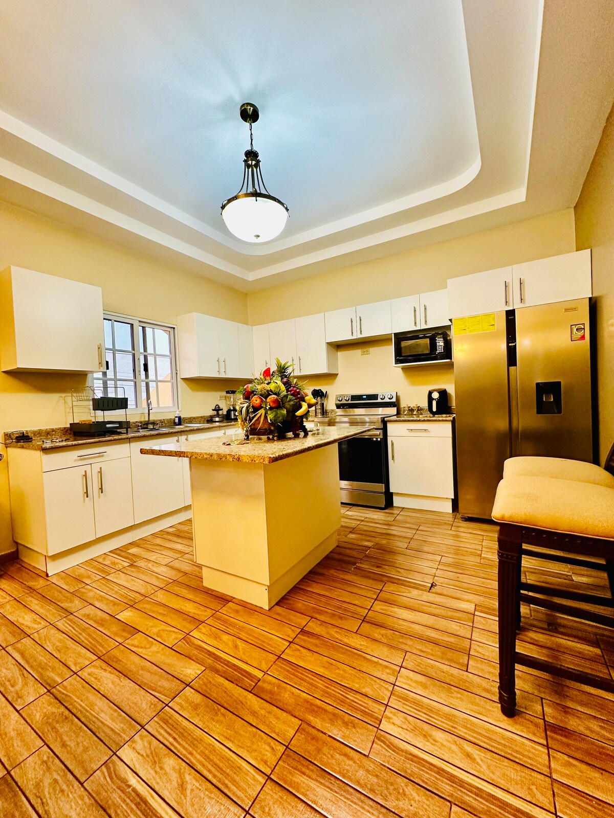 The bright kitchen features modern appliances, including a stainless steel refrigerator and oven. A central island offers additional counter space, complemented by a seating area with a high chair. Natural light streams through a window, illuminating the polished wooden floor.