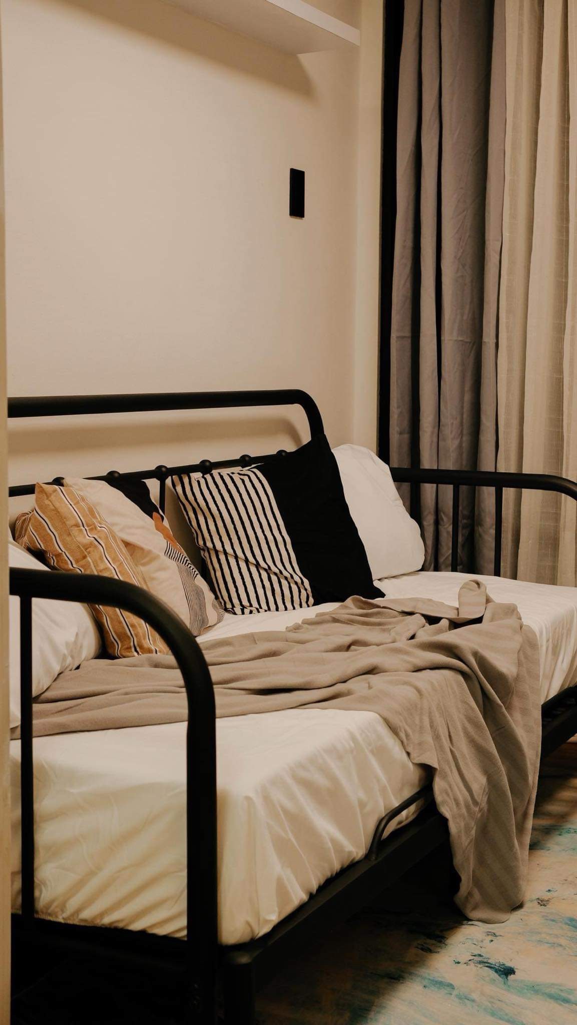 A versatile daybed is presented, with a neutral blanket draped casually over the edge. The bed is adorned with decorative cushions in various patterns. Soft, muted curtains frame the window, complementing the serene color palette of the room.