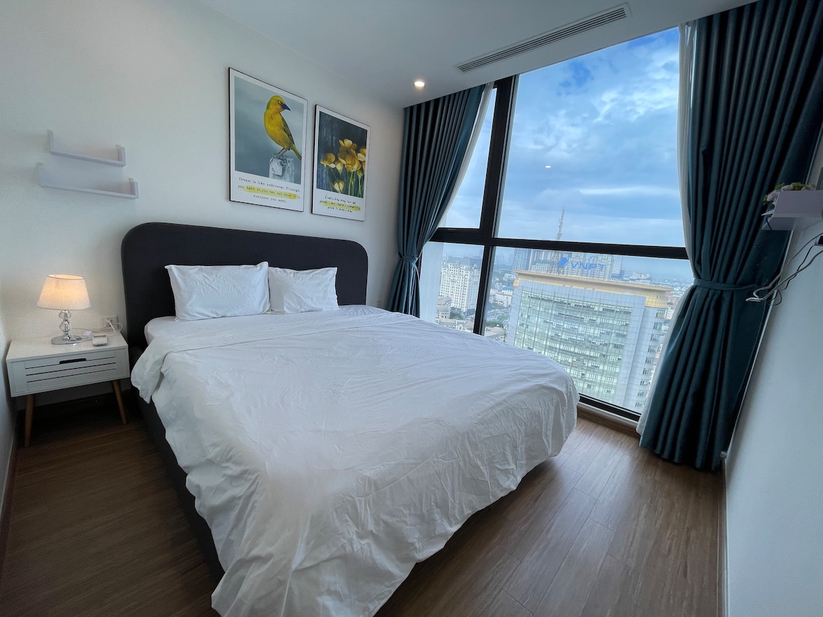 A bright bedroom features a spacious bed with crisp white linens. Artwork of birds and flowers adorns the wall. Large windows provide ample natural light and offer views of the city skyline, framed by neatly drawn curtains.