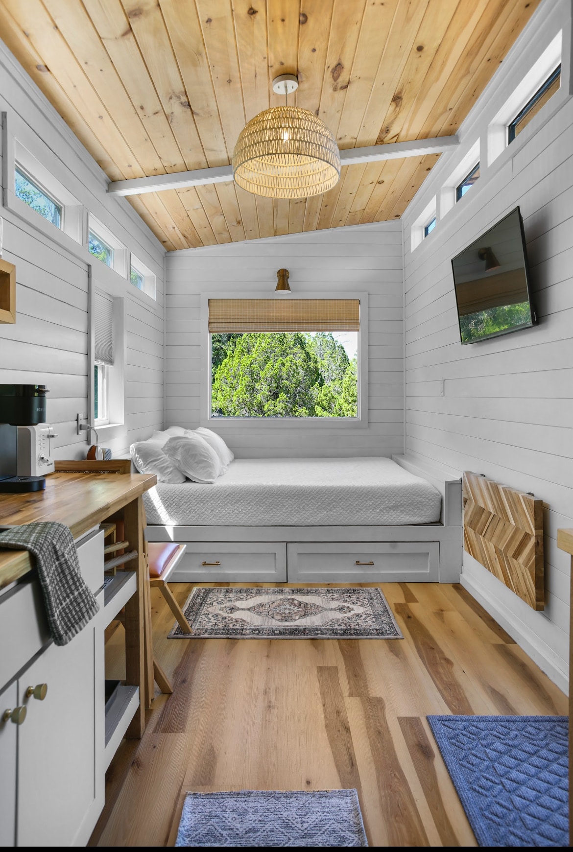 A cozy interior presents a spacious bed with soft linens, situated beneath a wooden ceiling. Natural light flows through the large window, showcasing views of greenery outside. A kitchenette with essential appliances and a small dining area is visible, complemented by warm wood flooring and decorative rugs.