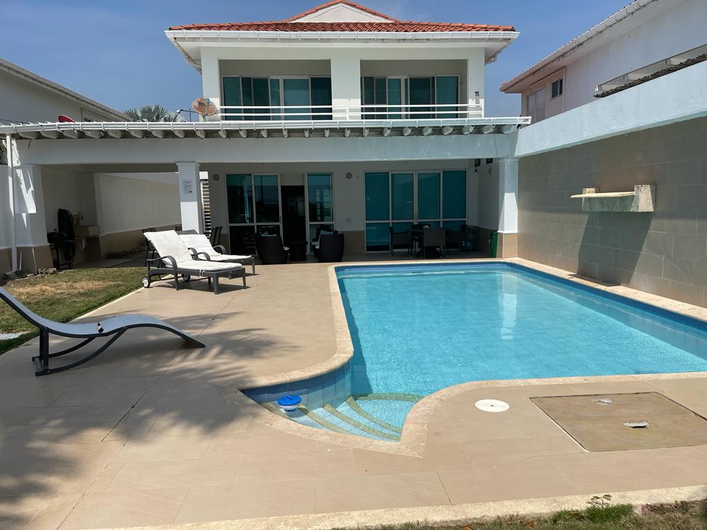 An outdoor area is depicted featuring a clear blue pool surrounded by a smooth tiled patio. Lounge chairs are arranged along one side, while a covered seating area adjoins the house. The exterior showcases large glass doors leading into the home.