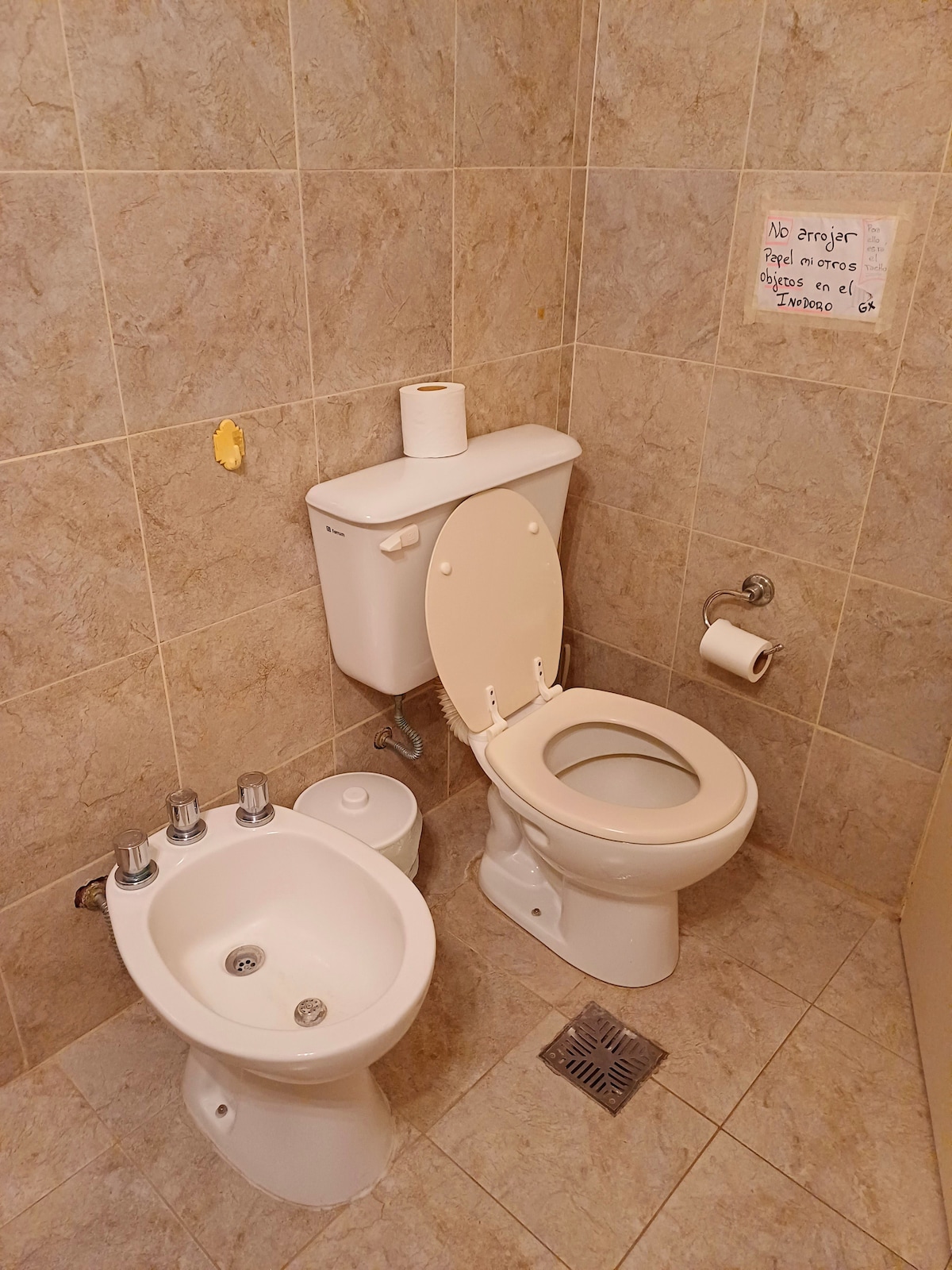 The bathroom features beige and gray tiled walls and floors. A standard toilet and bidet are positioned next to each other. A roll of toilet paper is placed on the tank. A wall sign offers instructions regarding disposal practices.