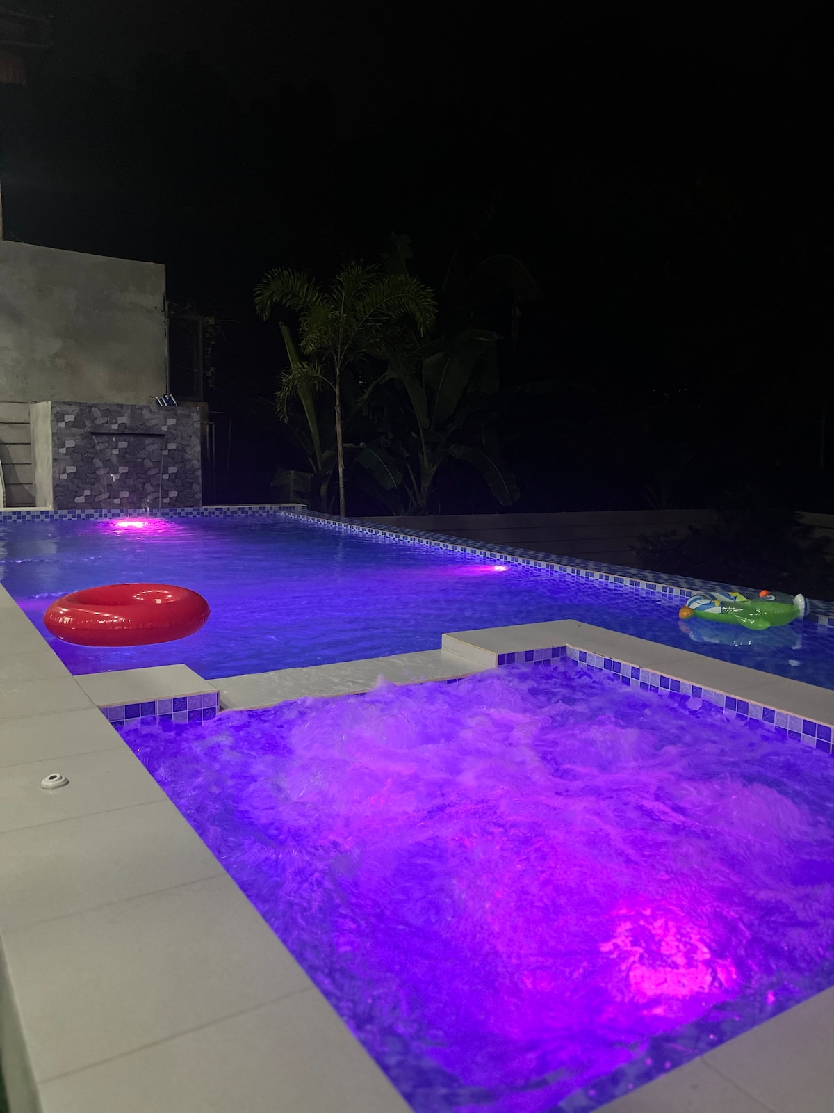 A tranquil pool area is showcased at night, featuring illuminated blue water and colorful light accents. A hot tub is included next to the pool, both surrounded by tropical vegetation. Floating toys, including a red ring and a green inflatable, add a playful touch.