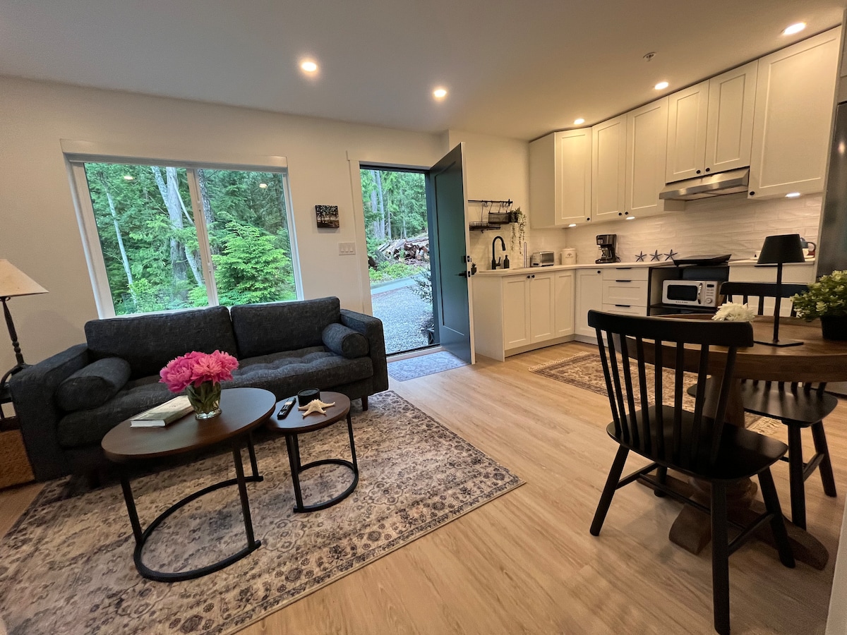 A bright living space features a comfortable couch beside a coffee table adorned with flowers. A kitchen area is equipped with modern appliances, while a dining table with chairs is situated nearby. Large windows overlook the lush greenery outside, inviting natural light into the room.