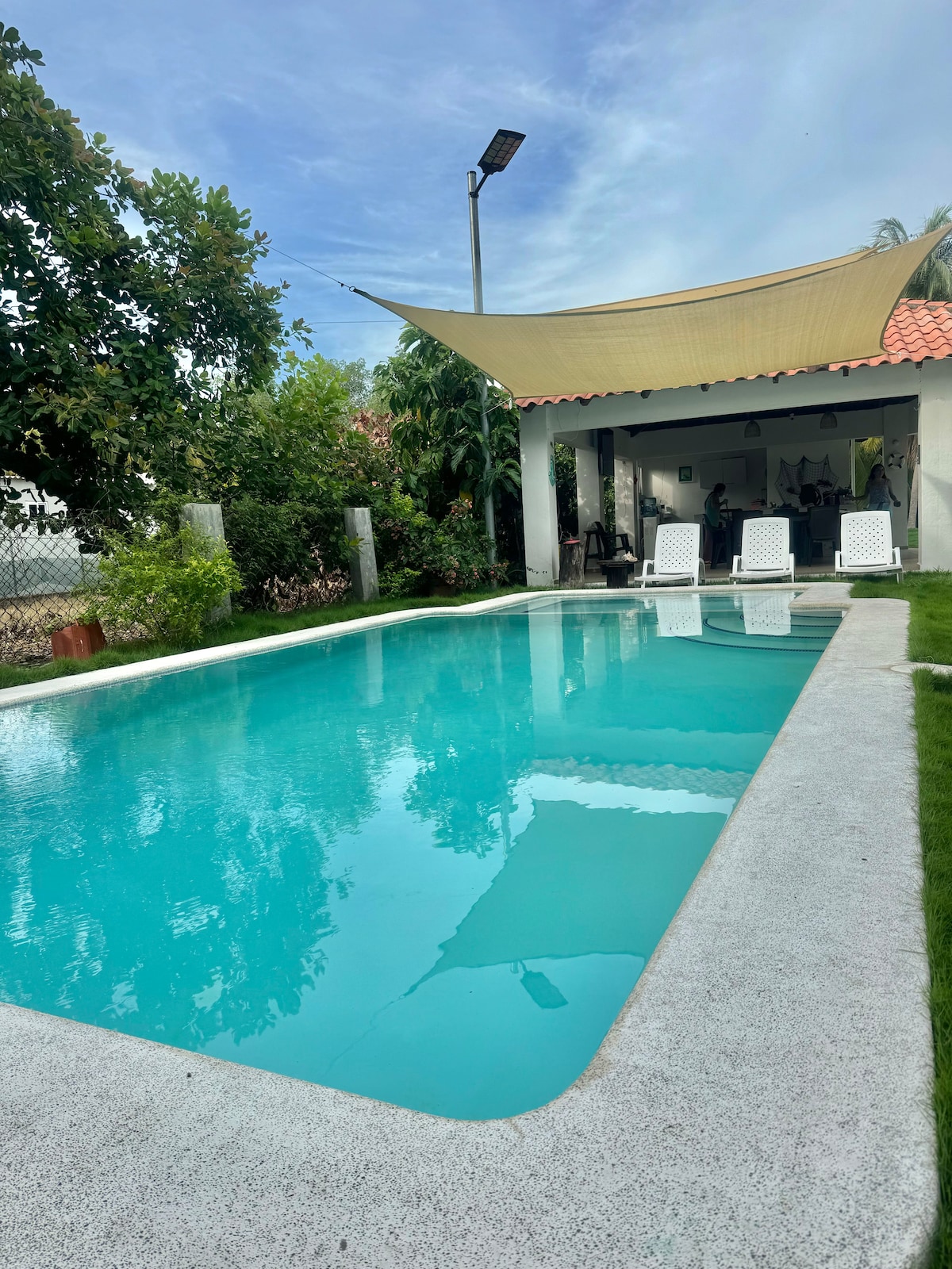 A clear, inviting pool is surrounded by lush greenery, with chaise lounges positioned for relaxation. A shaded patio space is visible in the background, offering a comfortable area for outdoor gatherings.