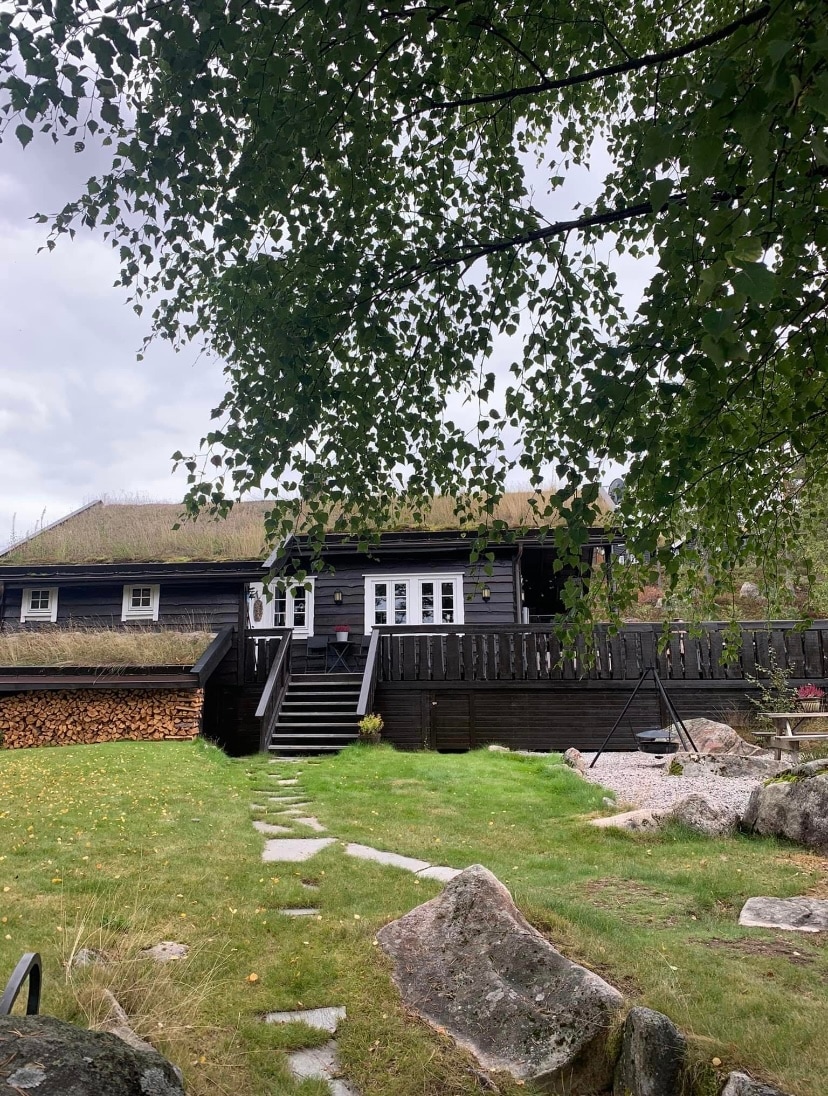 The image displays a charming cabin with dark timber siding, framed by lush greenery. Steps lead from the grassy yard to a spacious deck. A woodpile is neatly stacked beside the cabin, while large stones and pebbles are scattered across the outdoor space.