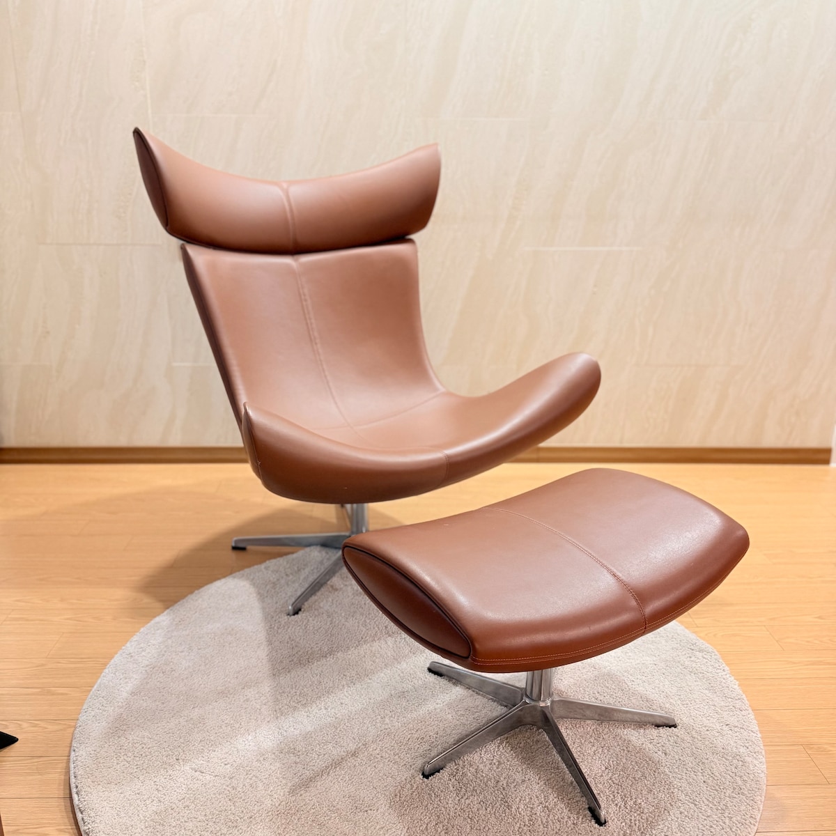 A modern armchair with a sleek, curved design and a matching footrest is displayed on a circular rug. The chair features a soft, tan leather upholstery, complemented by a polished metal base, providing a comfortable and stylish seating area.