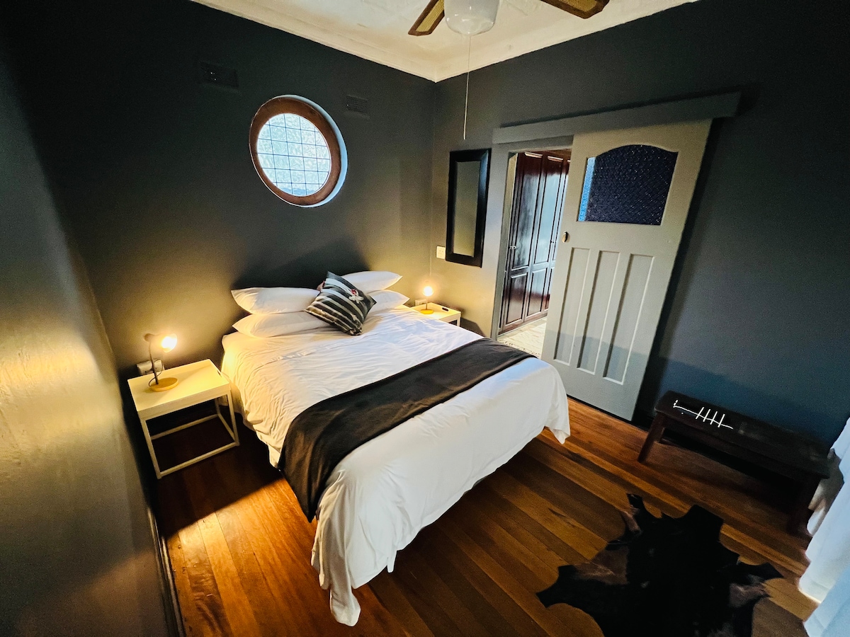 A cozy bedroom features a round window allowing natural light to greet a spacious bed adorned with simple bedding. Soft lighting from bedside lamps complements the dark walls and wooden flooring, while a mirror reflects the inviting atmosphere.