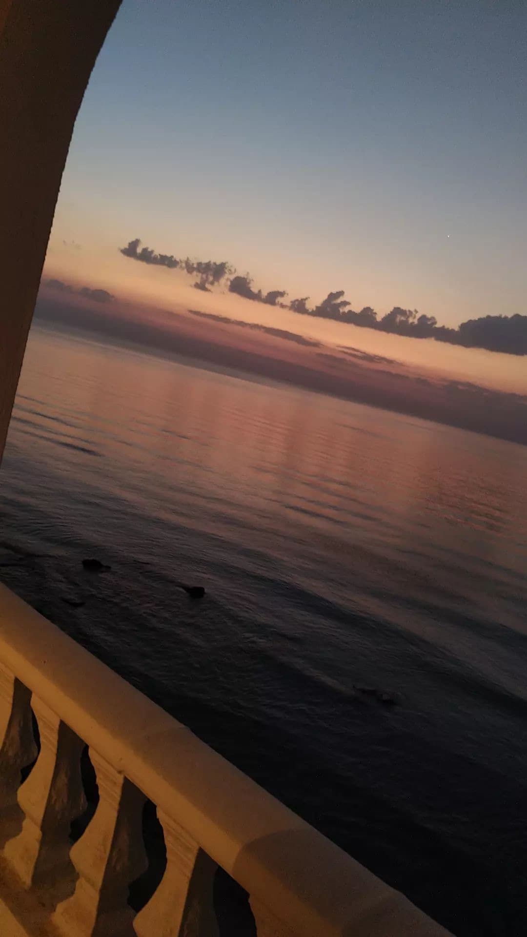 The image captures a serene seascape at sunset, featuring calm waters reflecting pastel shades of orange, pink, and purple. Wispy clouds stretch across the sky, and the horizon appears softened by the fading light. A portion of a balcony railing frames the scene.