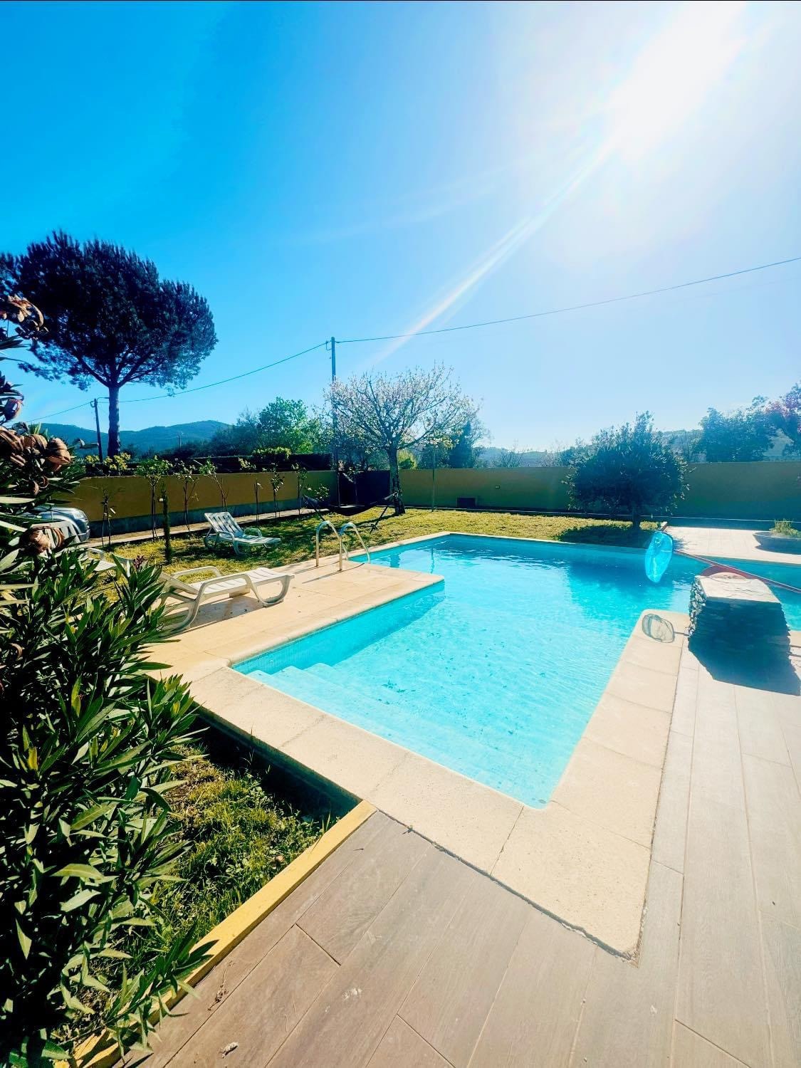 A clear pool glistens under the sun, surrounded by manicured greenery and trees. Sun loungers are positioned nearby on a spacious deck. The tranquil setting invites relaxation, with an unobstructed view of the blue sky above.