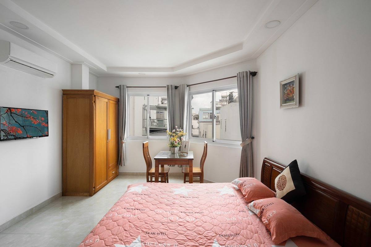 A well-lit bedroom features a spacious bed with a light pink quilt and decorative pillows. Large windows allow natural light to brighten the room, complemented by sheer curtains. A wooden wardrobe and a small dining table with chairs are positioned nearby, offering functional space.