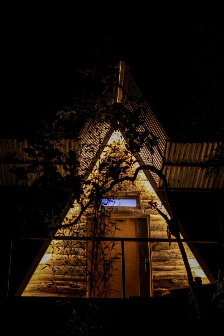 An A-frame cabin is illuminated against the night sky, highlighting its unique architectural shape. Soft light emanates from the entrance, framed by natural foliage, creating a serene and inviting atmosphere for guests.