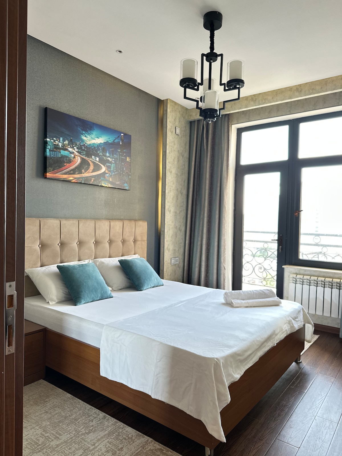 The bedroom features a spacious bed with soft linens and decorative pillows. Large windows allow natural light to fill the room, while a striking cityscape painting adds a modern touch. A tasteful light fixture enhances the ambiance, contributing to a comfortable environment.