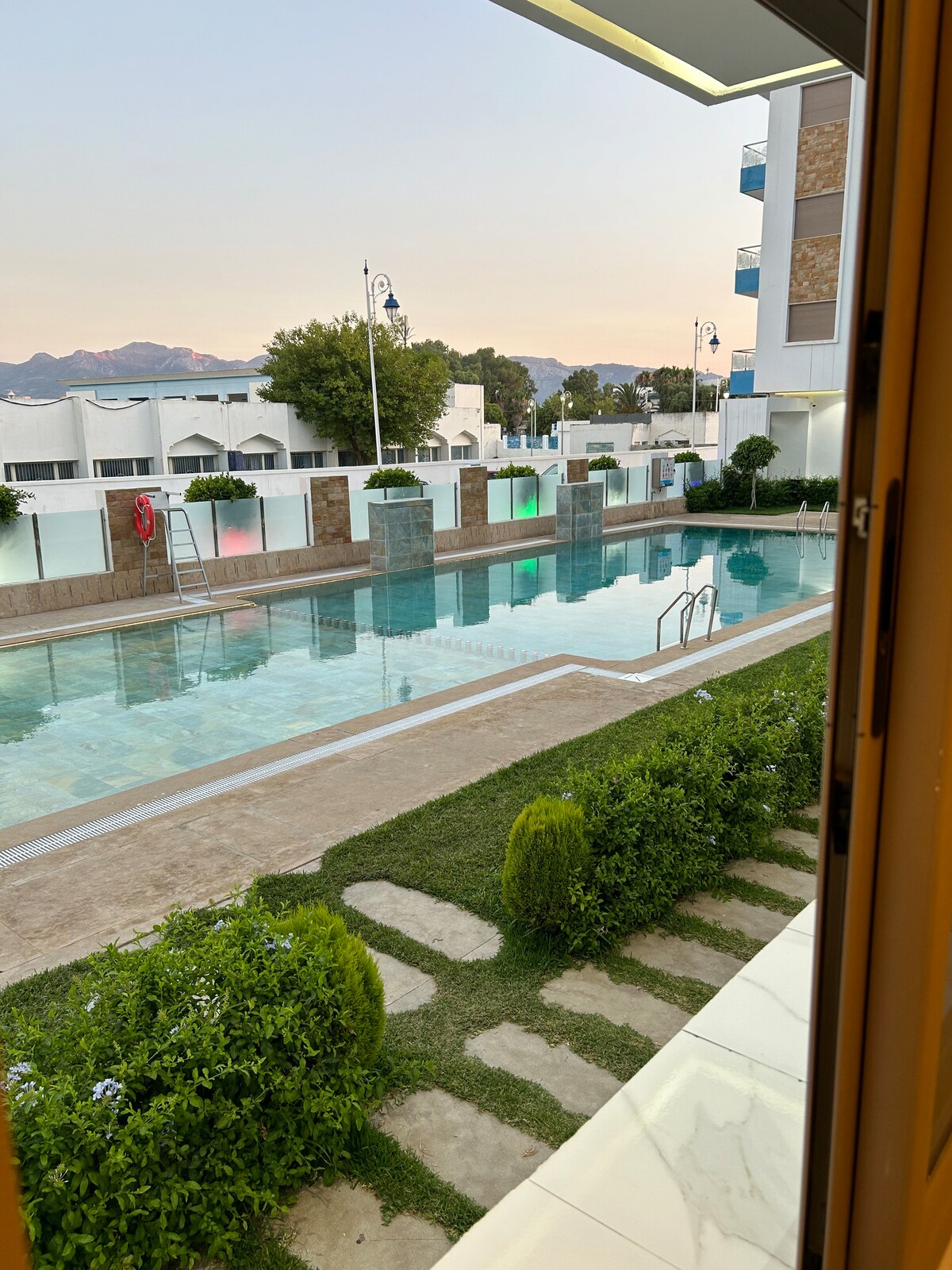 A view of the outdoor swimming pool is presented, featuring clear water and surrounding greenery. The pool is framed by smooth stone pathways and well-maintained grass. Modern buildings are visible in the background, with soft evening light casting a warm glow over the scene.