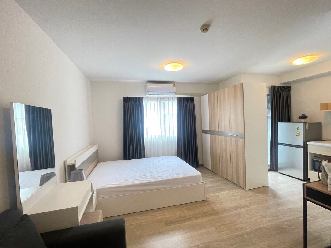 A cozy bedroom is presented, featuring a spacious bed without bedding. A large mirror and a white storage unit enhance the room's functionality. A window with sheer curtains allows natural light, while a compact refrigerator and a dining area with a table are visible in the background.