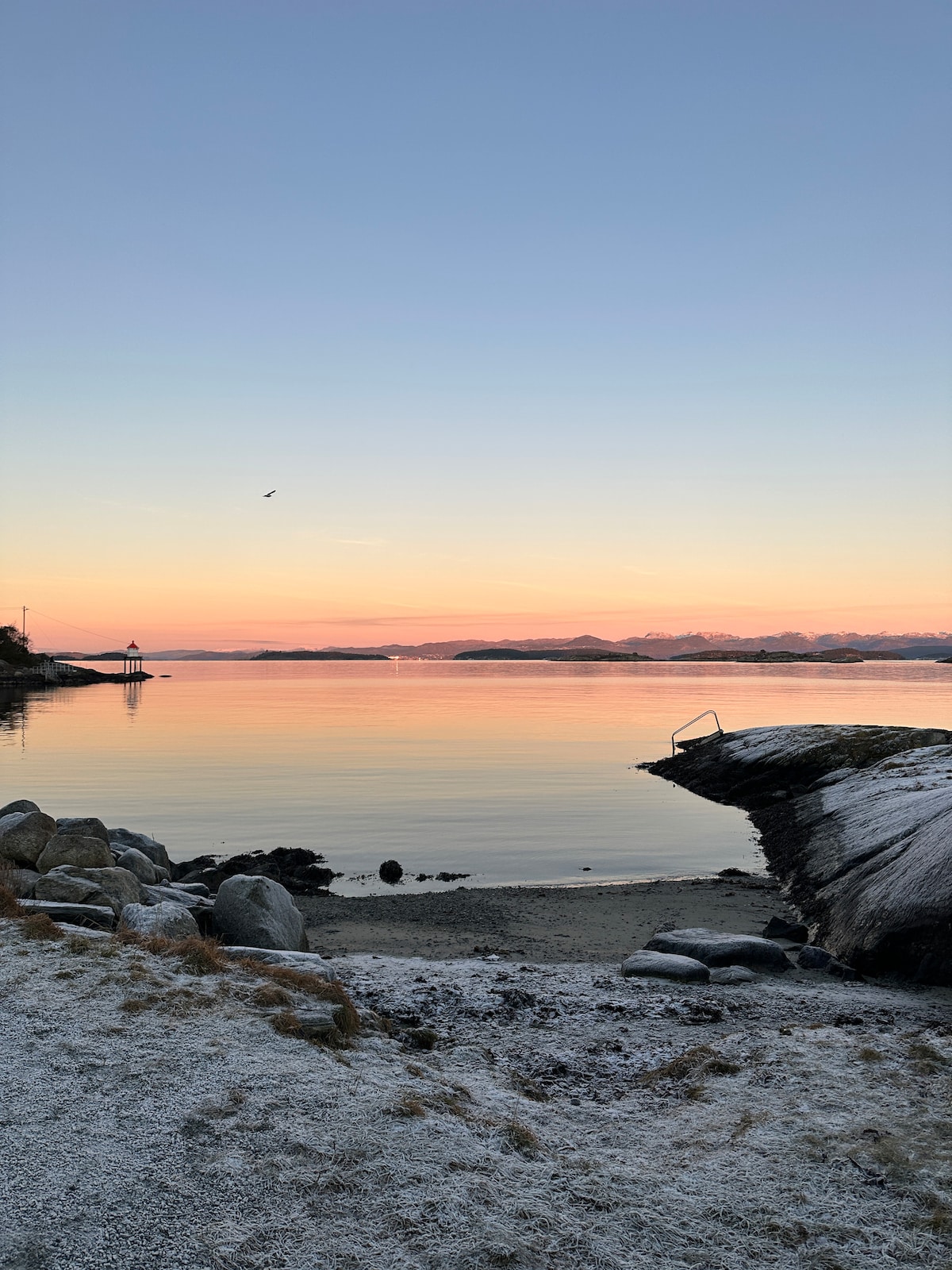 A calm water scene is captured at sunset, with pastel hues reflecting off the surface. Smooth stones line the shore, leading to a wooden structure and gentle waves lapping at the edge. The distant outline of hills is visible under a clear sky.