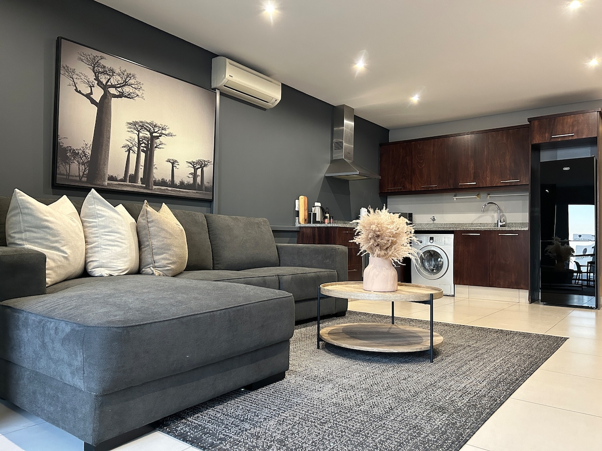A contemporary living area is highlighted by a dark grey sectional sofa adorned with plush pillows. A round coffee table stands atop a textured rug, positioned centrally. The kitchen features dark wood cabinetry and modern appliances, with a washing machine discreetly integrated into the design.