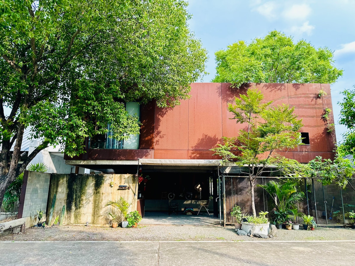 The exterior of the house is showcased, featuring a combination of rust-colored metal and green foliage. Large windows allow natural light to enter the living spaces. The entrance is marked by a covered area, surrounded by plants, creating an inviting ambiance.