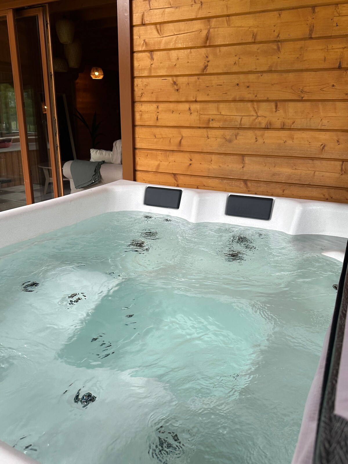 A private hot tub is set beneath a wooden terrace, surrounded by a cozy cabin atmosphere. The water is clear, inviting relaxation, with strategically positioned jets visible along the bottom. Soft lighting from the cabin enhances the tranquil setting.