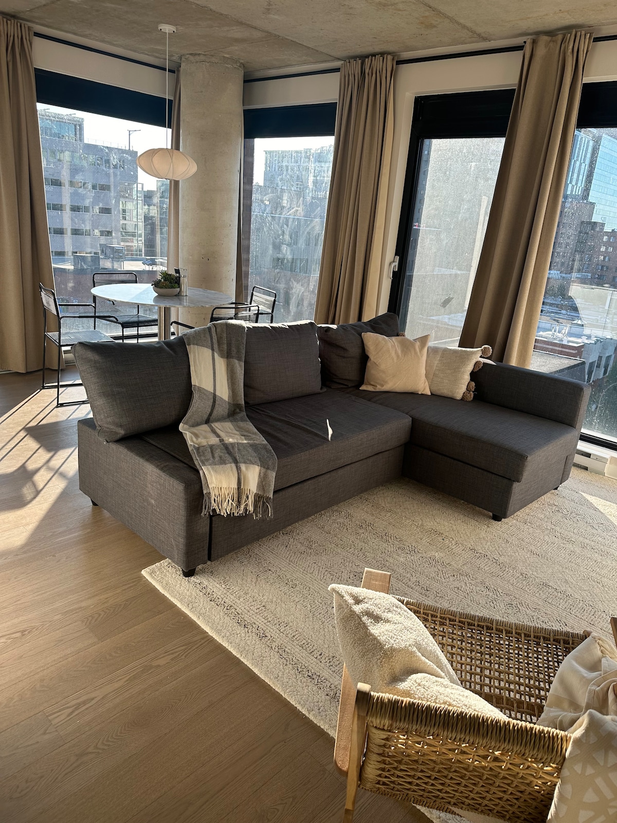 A corner seating area is shown, featuring a dark sectional sofa with a beige throw blanket and decorative cushions. Natural light streams in through large windows, illuminating a modest dining table with black chairs. Beige curtains frame the glass, enhancing the bright interior.