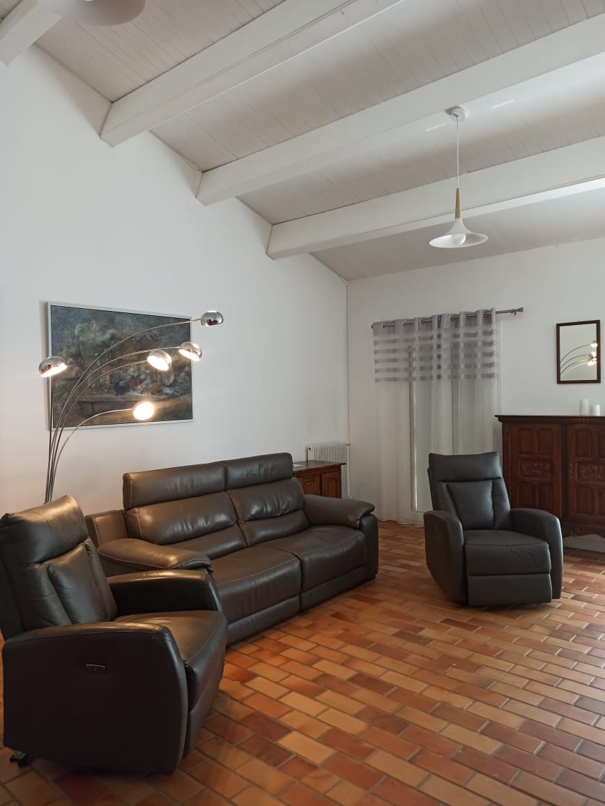A living area features two dark leather seating options, including a reclining chair. Natural light enters through a window covered with sheer curtains. A textured artwork enriches the wall above the seating, and a wooden cabinet offers additional storage space.