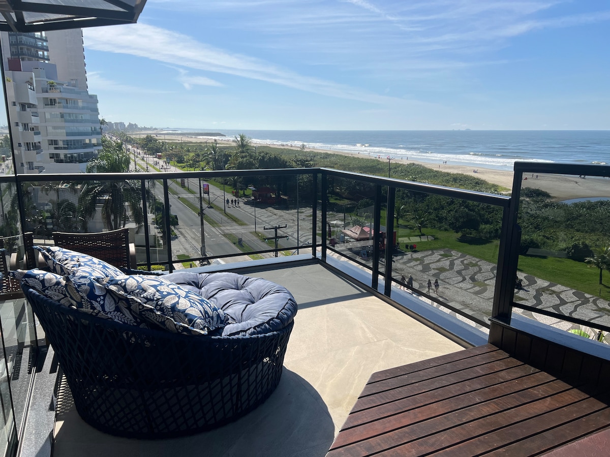 A spacious balcony features comfortable seating with plush cushions, offering unobstructed views of the beach and ocean. The glass railing enhances the panoramic vista, while sunlight illuminates the area, highlighting the inviting outdoor atmosphere.