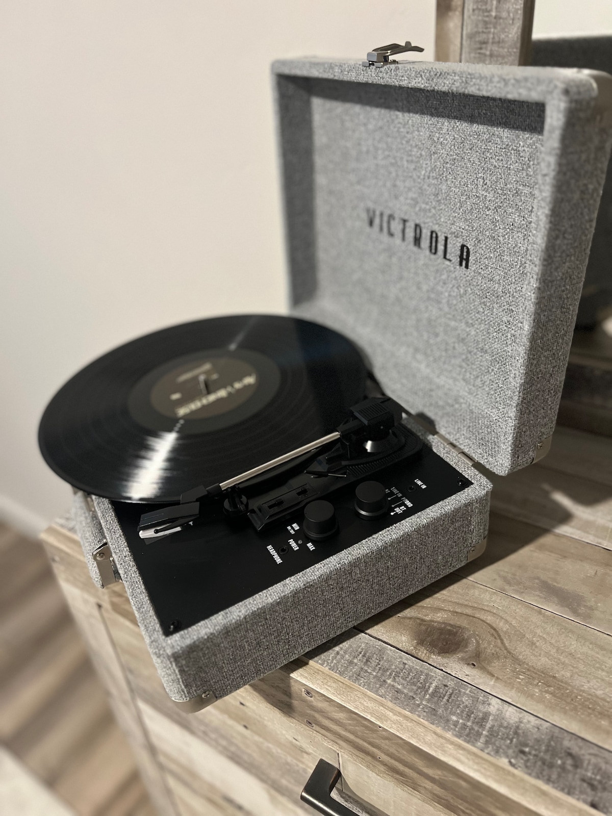 A vintage-style record player is set on a wooden surface, displaying a vinyl record on its turntable. The player is housed in a grey case with a textured finish, featuring simple controls for an effortless listening experience.