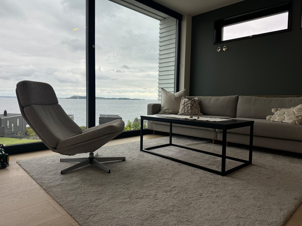 A comfortable living area features a sleek gray sofa and a modern armchair, positioned on a soft rug. A coffee table is centered in front, while large windows frame a panoramic view of the water, inviting natural light into the space.