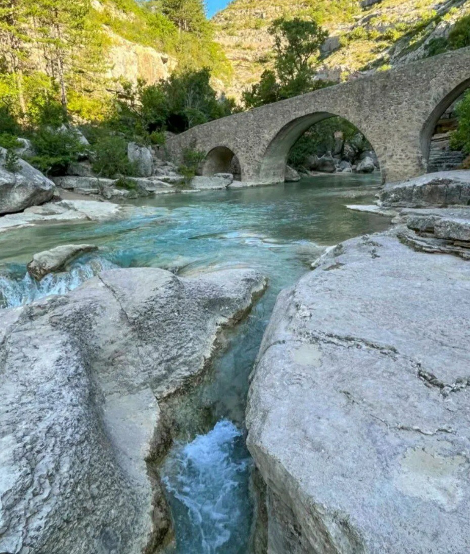 A gentle river flows through a rocky landscape, with clear turquoise water reflecting the surrounding greenery. An arched stone bridge spans the water, adding a historical element to the serene natural scene. Boulders and smooth stones line the riverbank.