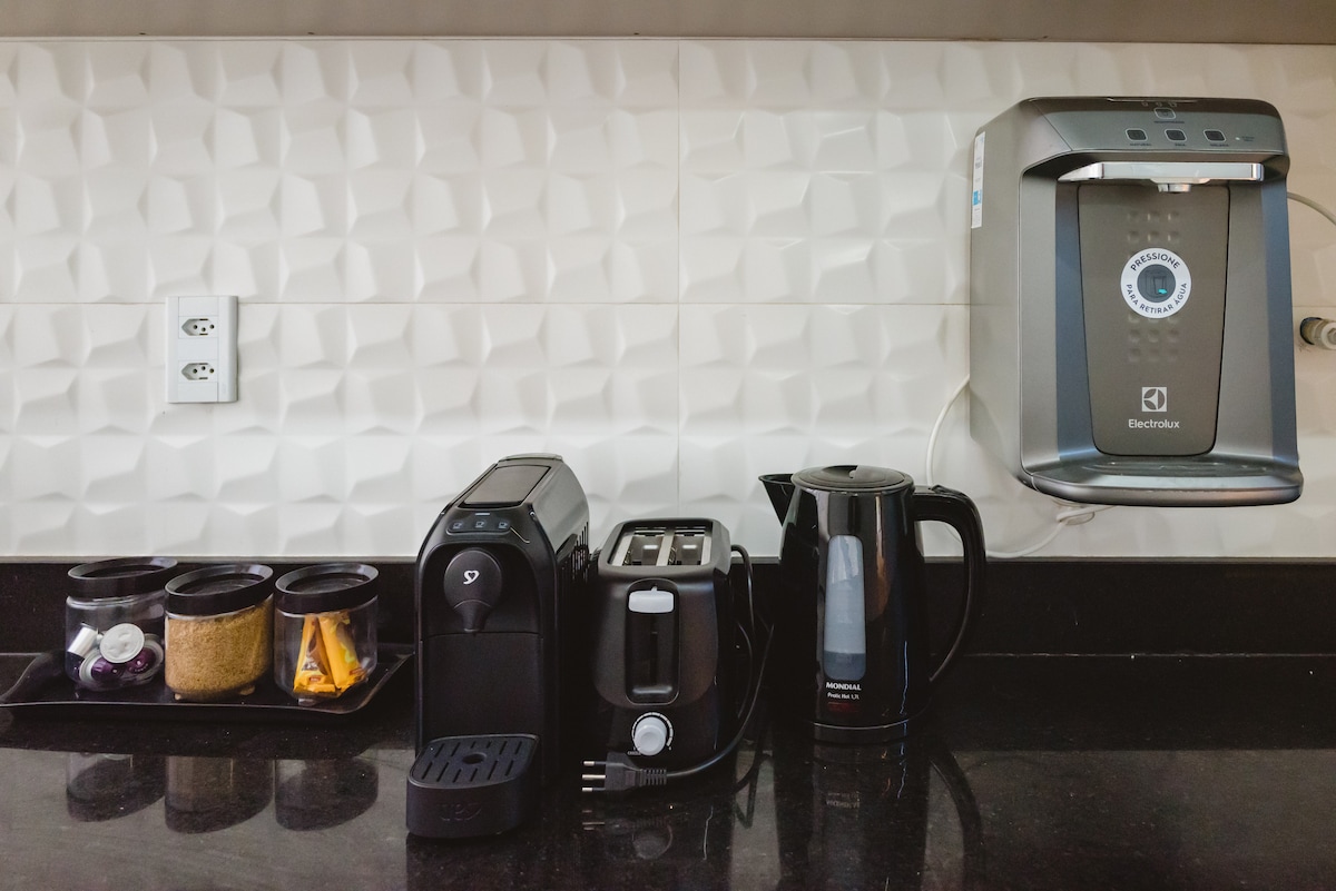 A modern kitchen countertop displays various appliances, including an espresso machine, toaster, electric kettle, and hot water dispenser. Containers for ingredients are arranged nearby, complementing the clean lines of the textured backsplash. The countertop features a dark finish, enhancing the room's sleek appearance.