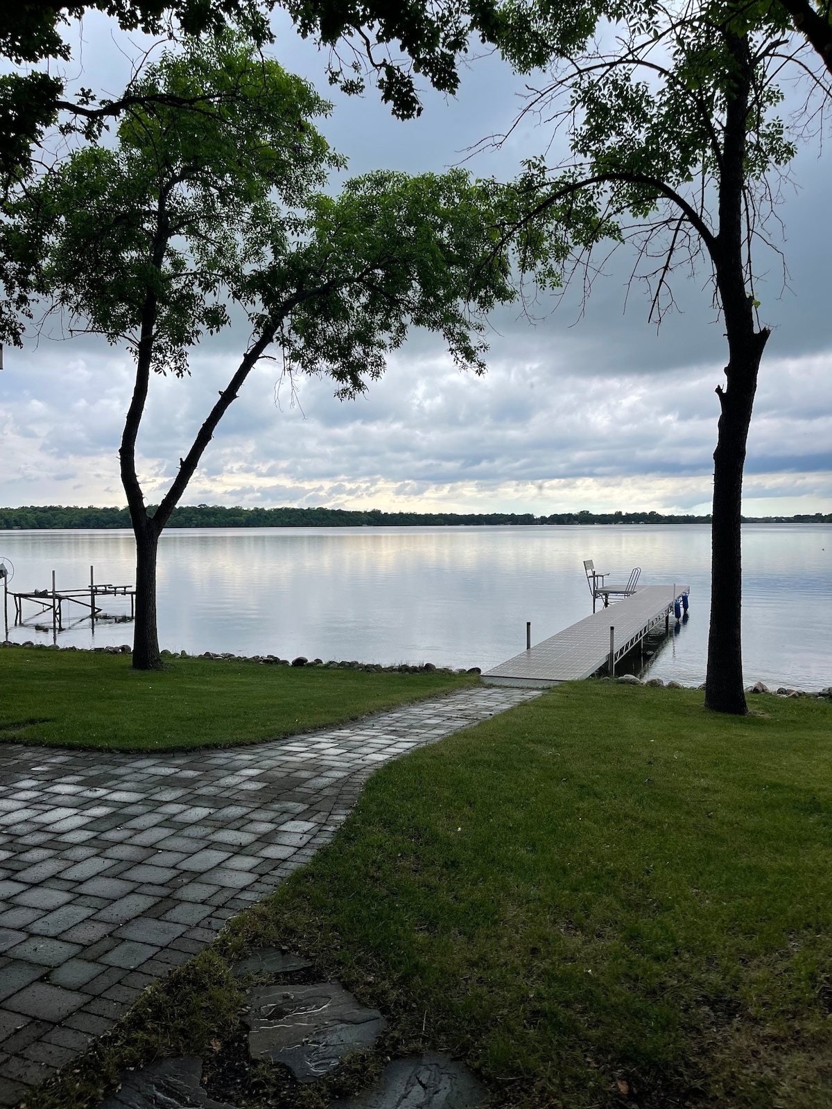 A stone pathway leads to a wooden dock extending over calm waters, framed by lush green trees. The sky is overcast, creating a serene atmosphere. Reflections of the trees can be seen in the water, enhancing the tranquil setting.