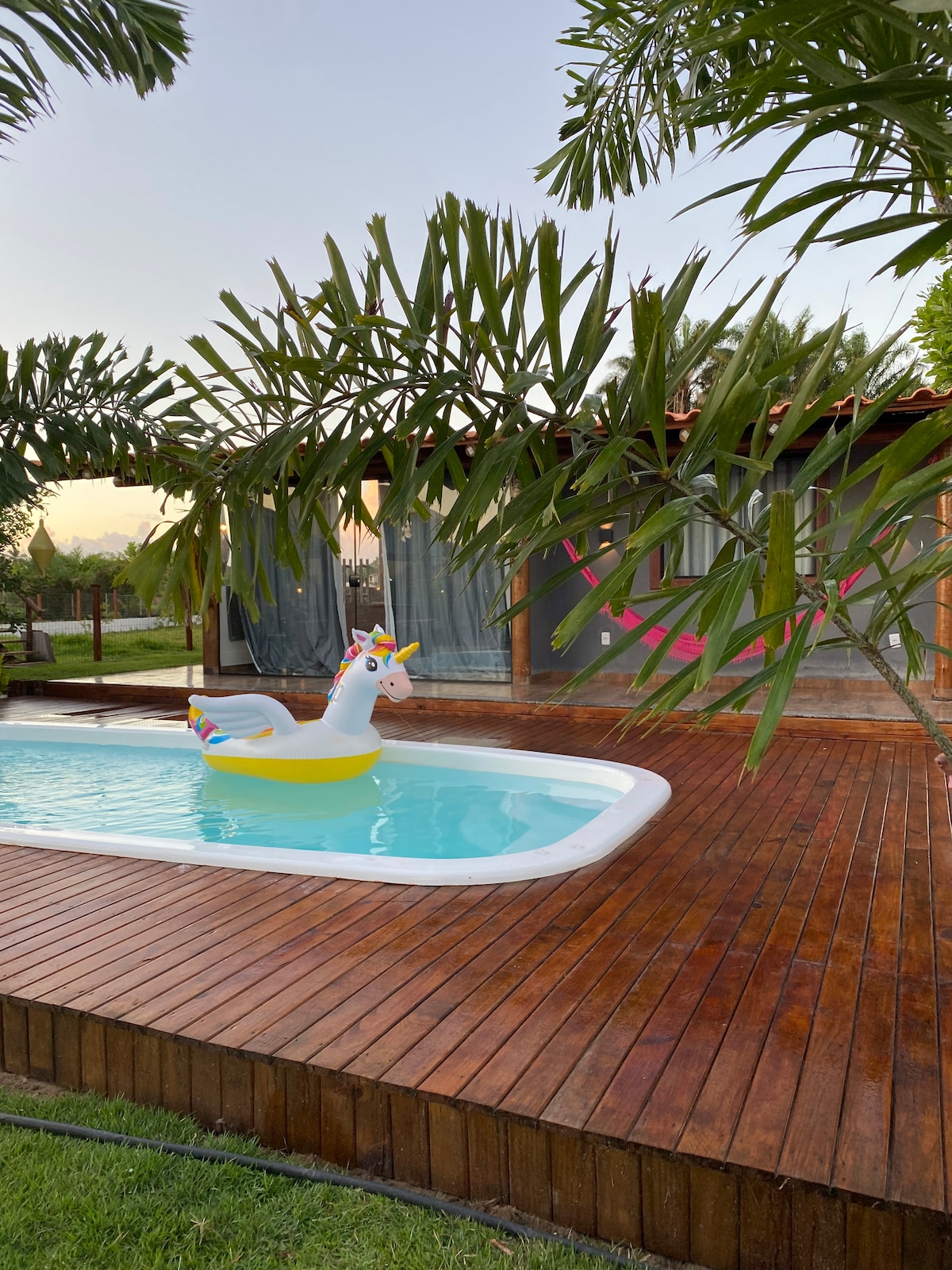 A clear swimming pool features a colorful unicorn float, situated on a wooden deck surrounded by lush greenery and palm trees. A rustic structure with large windows is visible in the background, suggesting a serene outdoor space.