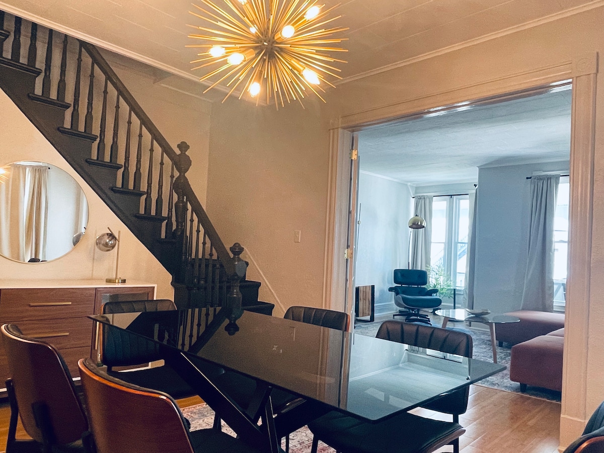 A modern dining area is presented with a sleek black glass table surrounded by six chairs. A unique starburst chandelier hangs overhead, illuminating the space. A staircase is visible in the background, leading to an upper level, while a glimpse of a cozy living room can be seen through an open doorway.
