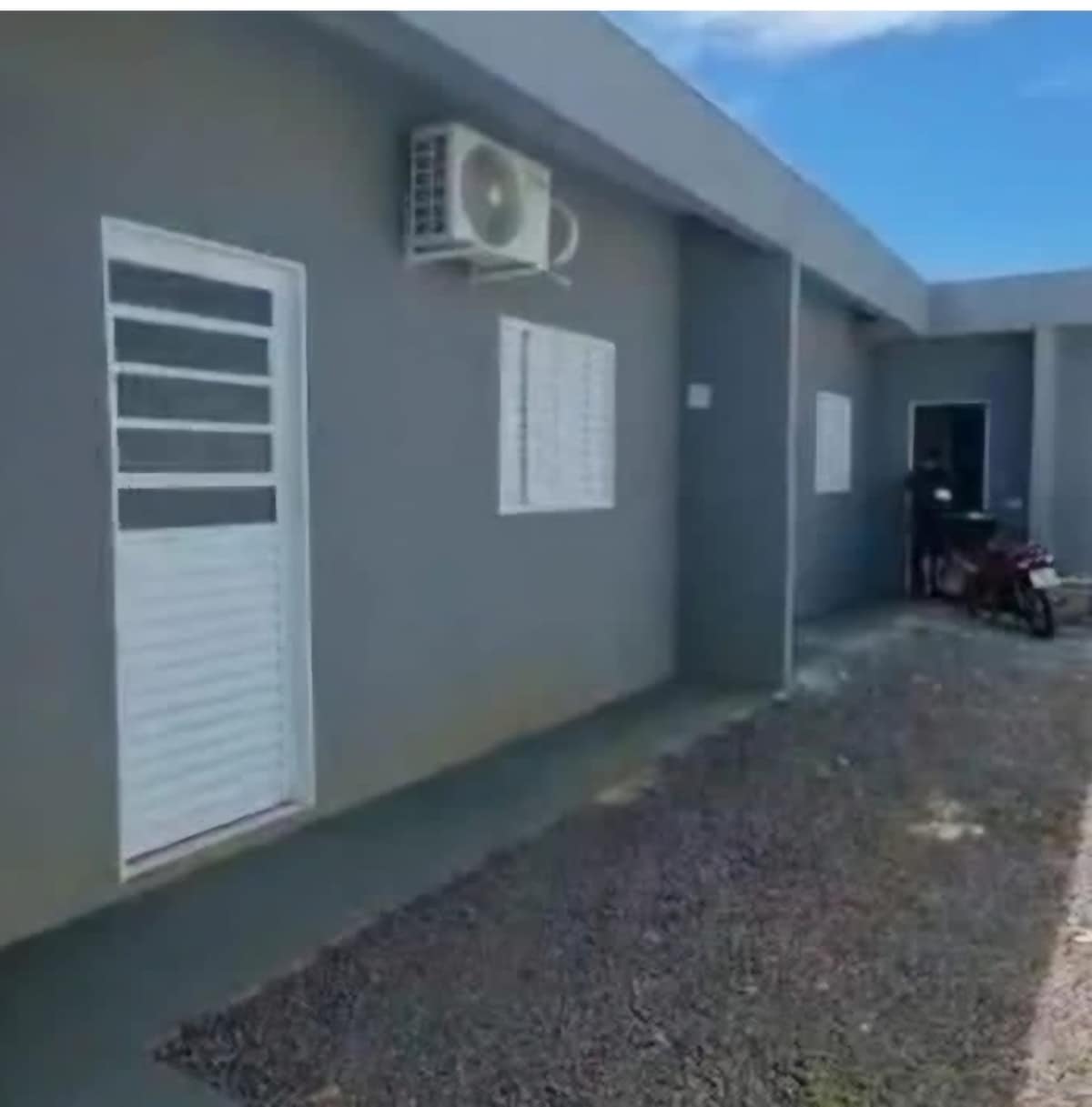 The exterior view features a modern building composed of gray walls, complemented by a white door and window. An air conditioning unit is visible above the door, while a small pathway lined with gravel leads to the entrance, creating a clean and accessible approach.