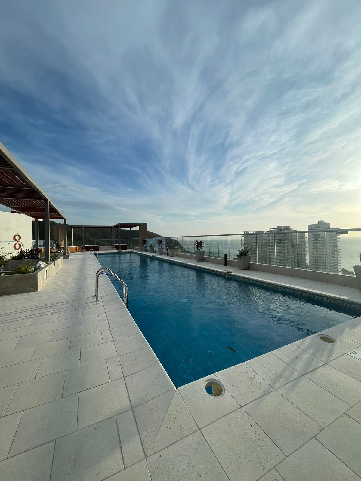 A rooftop pool area is presented with clear blue water reflecting the sky. Surrounding the pool, lounge chairs are arranged on a spacious deck. A panoramic view of city buildings and the horizon can be seen in the distance under a partly cloudy sky.