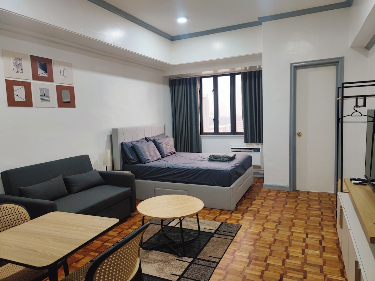 The studio unit features a cozy sleeping area with a spacious bed dressed in soft linens. A comfortable sofa sits nearby, complemented by a coffee table on a patterned rug. Natural light filters through the window, with curtains drawn for privacy.
