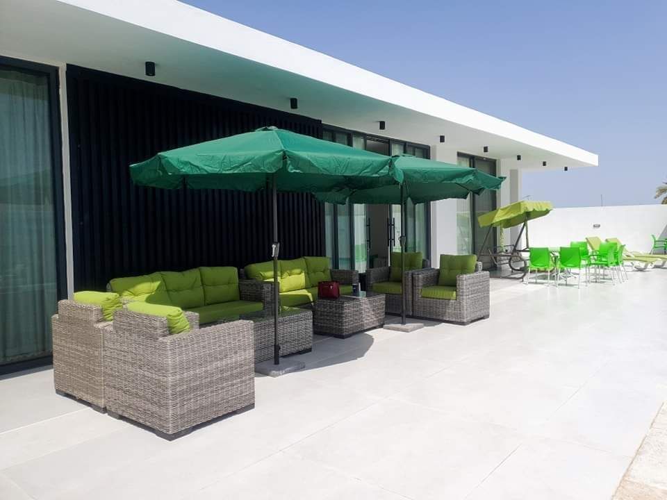 A shaded outdoor seating area is furnished with green umbrellas and inviting couches. The furniture features a combination of light-colored cushions and woven frames. Large glass doors provide an open feel, connecting the space to the surrounding pool area.