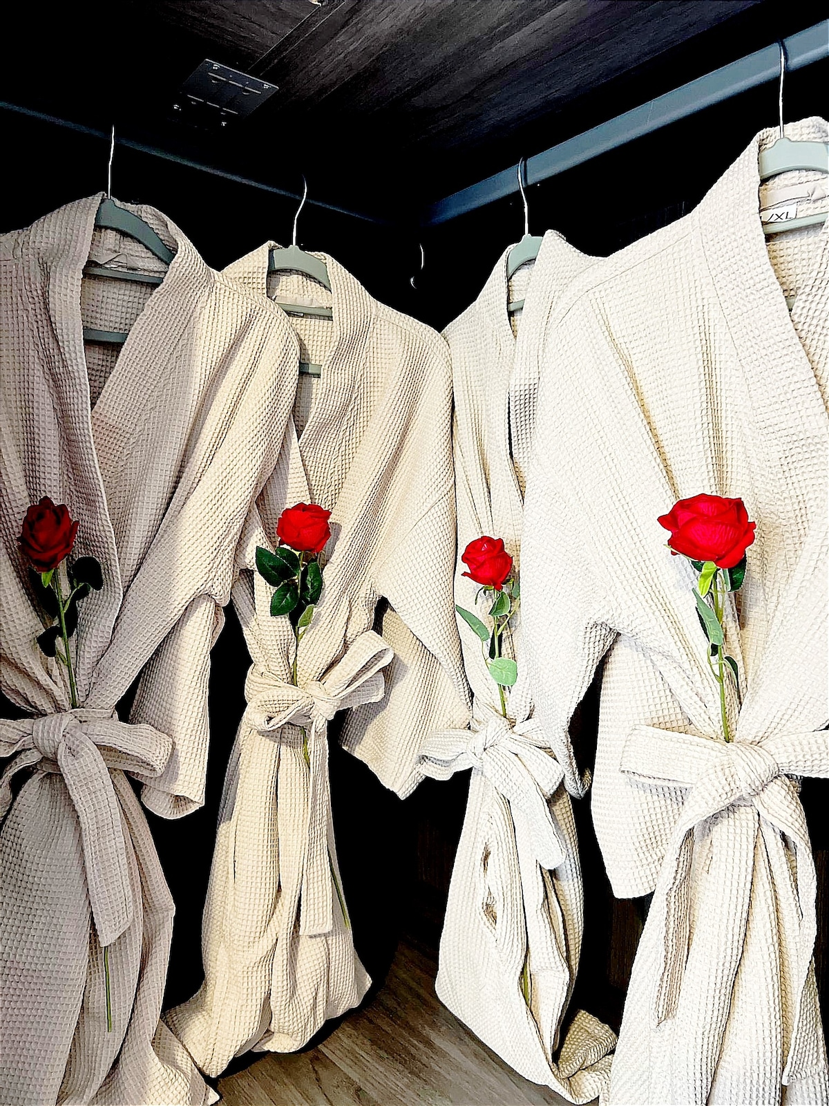 Four beige bathrobes are displayed on hangers, each adorned with a red rose placed delicately in the fold. The robes feature a textured pattern and are tied at the waist, creating a serene and inviting presentation in the closet.