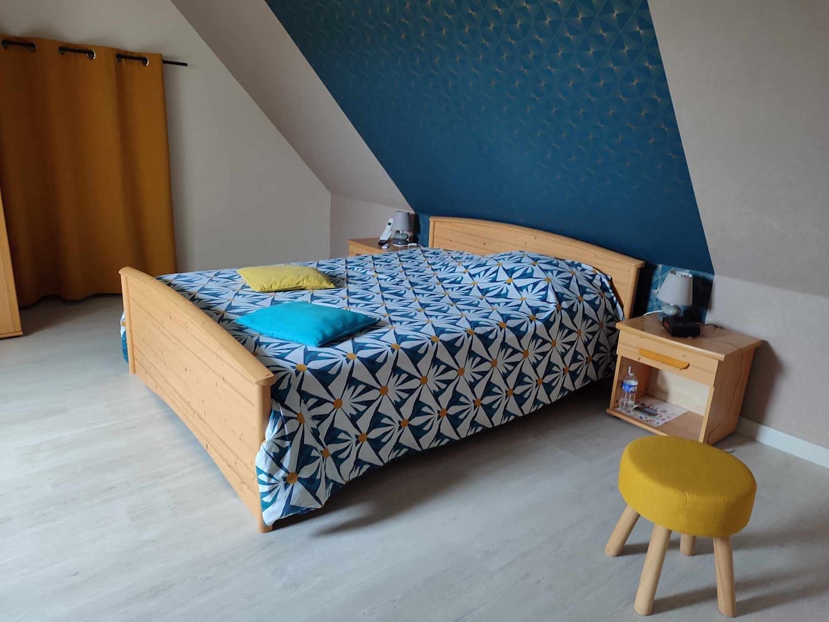 A bright sleeping area features a double bed adorned with a patterned blanket. Flanking the bed are two nightstands, each accompanied by a lamp. A stool adds a pop of color, while a curtain in warm tones partially covers the window, enhancing the room's coziness.