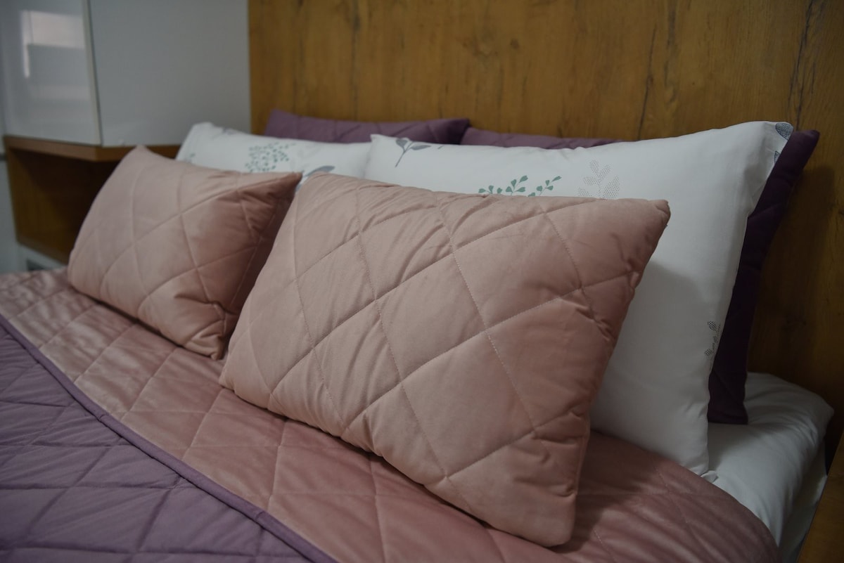 A cozy sleeping area is presented, featuring plush pillows and a soft quilt in complementary shades of pink and purple. The bedding is neatly arranged, with decorative pillows adding texture against a warm wooden backdrop.