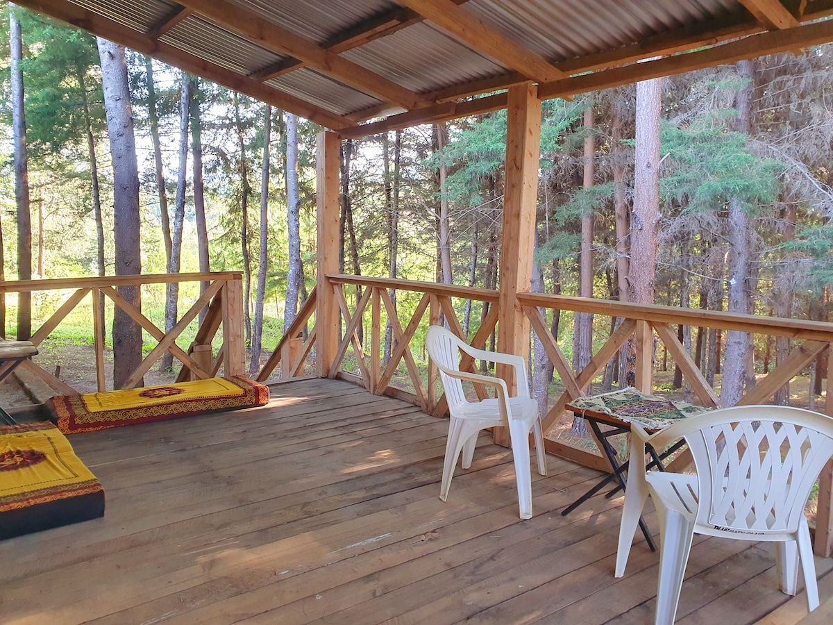 A covered porch is surrounded by trees, featuring a wooden floor and a railing. Two mats are placed on one side, while two white chairs and a small table are arranged on the other. Natural light filters in, creating a peaceful outdoor setting.
