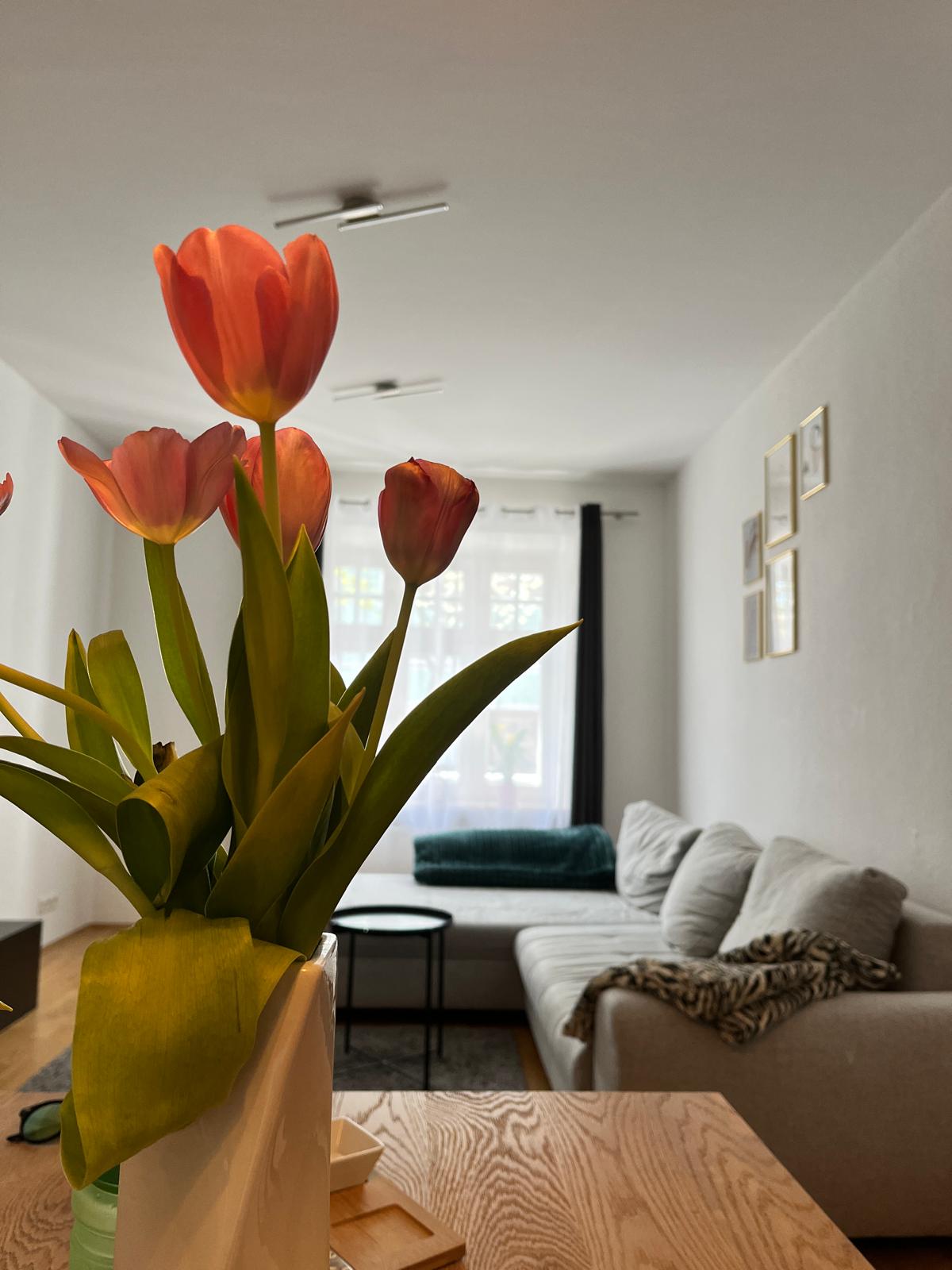A vase filled with vibrant tulips stands prominently in the foreground. In the background, a cozy seating area features a light-colored sofa, complemented by a dark side table. Soft natural light filters through a window, illuminating the space and highlighting the neutral color palette.