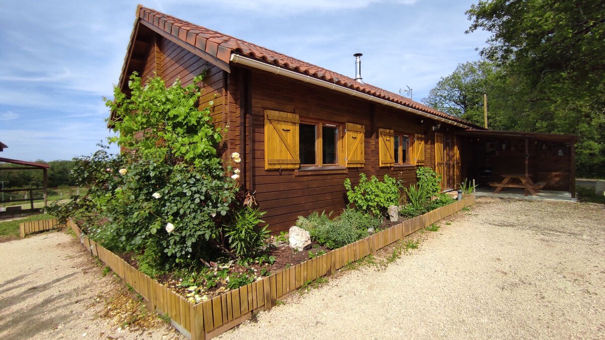 The exterior of a single-story wooden home is surrounded by a well-maintained garden. Yellow flowers and lush greenery are visible along the path. The house features brown shutters on the windows and a gravel driveway leading to the property.