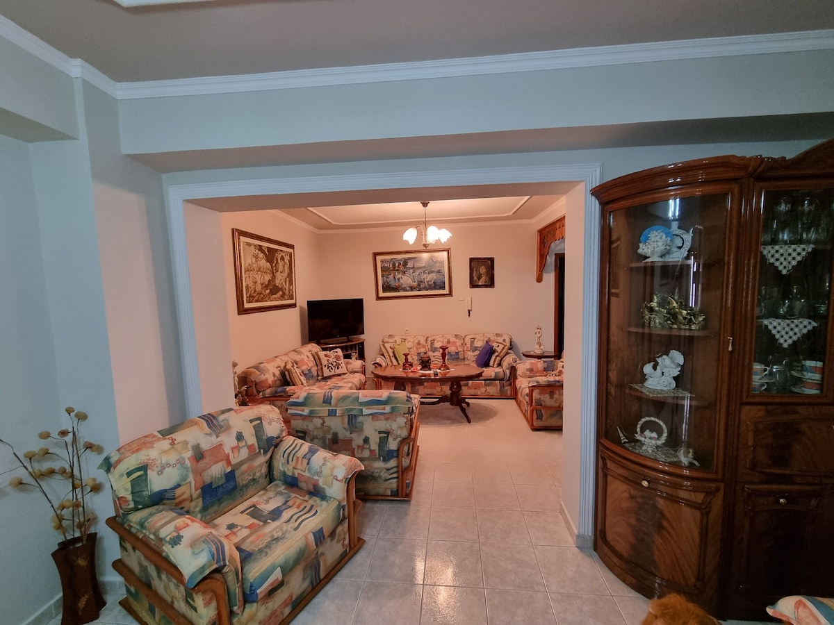 A comfortable living area features multiple patterned sofas arranged around a central table. A television is positioned on a stand against the wall. Decorative artwork and a cabinet filled with china add character to the space, while soft lighting creates a welcoming ambiance.