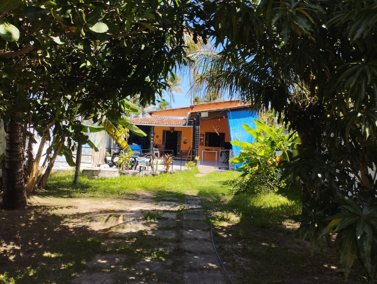 A vibrant accommodation is framed by lush greenery, with a pathway leading to the entrance. The exterior features a blend of blue and orange hues, while outdoor seating areas are visible under palm trees, inviting relaxation in a tranquil setting.