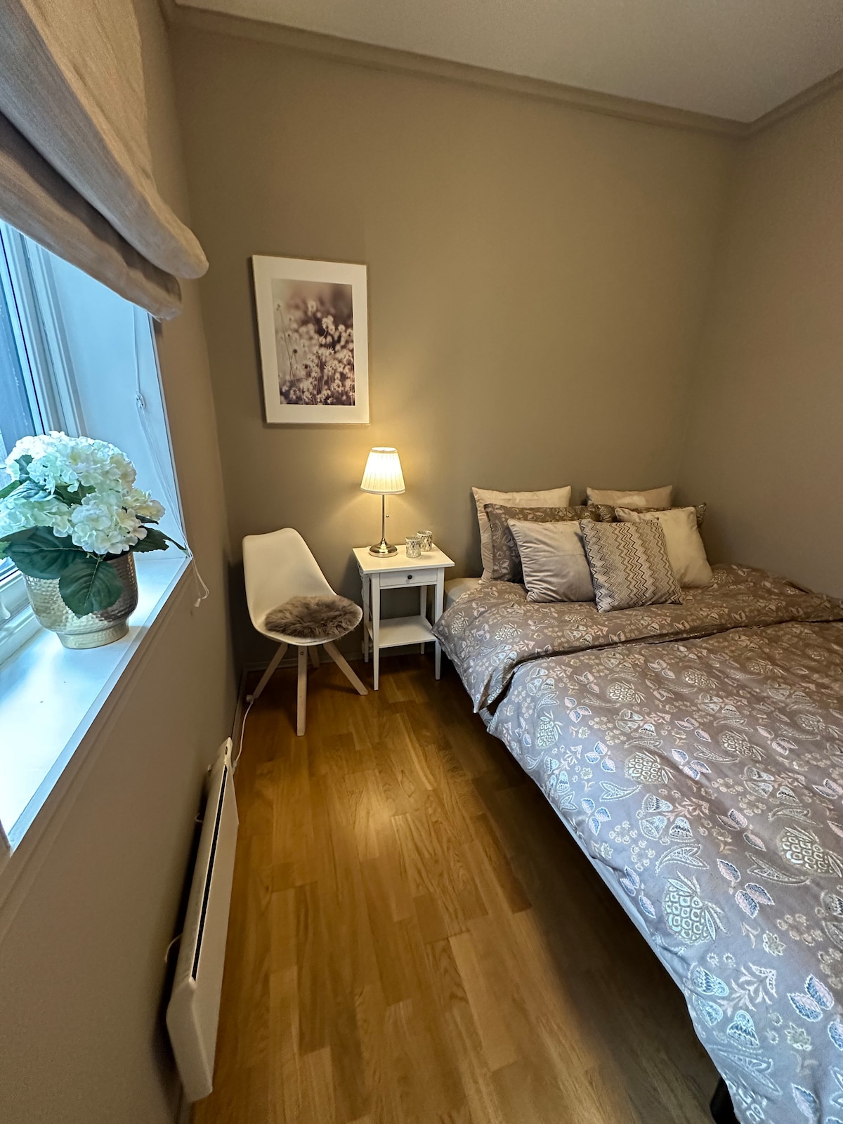 A well-appointed bedroom features a spacious bed draped in intricate bedding. A bedside table holds a lamp and decorative items, while a soft chair complements the space. Natural light enters through the window, accompanied by a small potted plant on the sill.