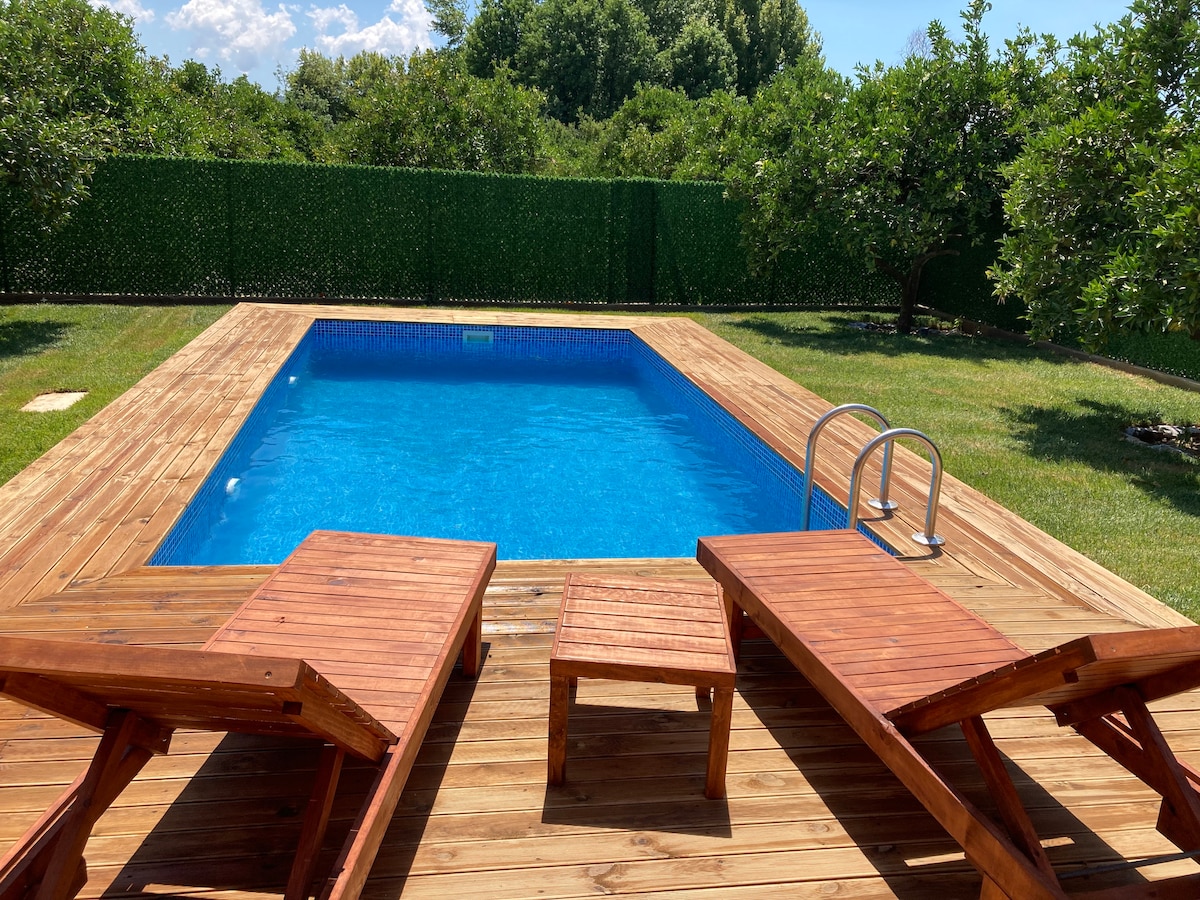 A private pool area is surrounded by lush greenery, featuring wooden decking. Two lounge chairs are positioned nearby, inviting relaxation. The pool is a clear blue, reflecting the sunlight, and steps lead down into the water, creating a welcoming outdoor space.