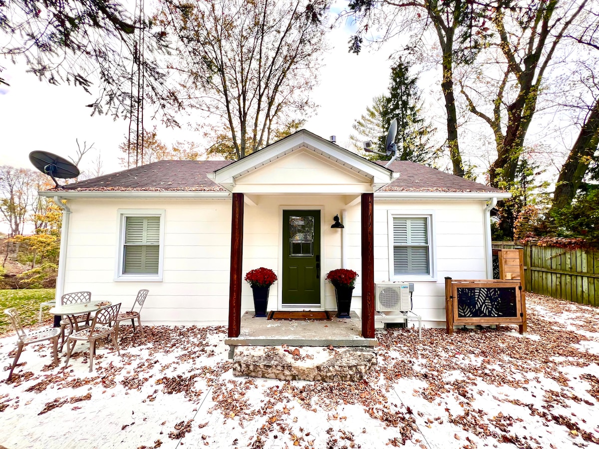 The charming exterior of a single-story house is surrounded by trees. A welcoming front porch is adorned with potted plants and a decorative screen. Two small chairs with a table are positioned on the porch, and leaves are scattered across the ground.