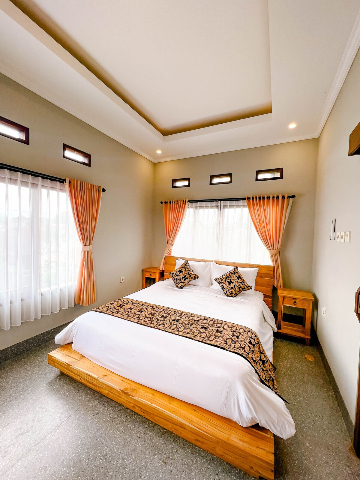 A bright sleeping area features a spacious bed adorned with a decorative blanket. Two bedside tables frame the bed, while natural light filters through sheer curtains and small windows, creating a serene ambiance complemented by soft grey walls.